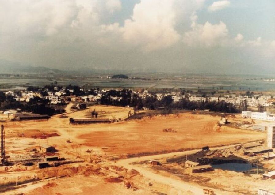 Aerial view of a construction site surrounded by low buildings and fields, indicating urban development amidst a rural landscape.