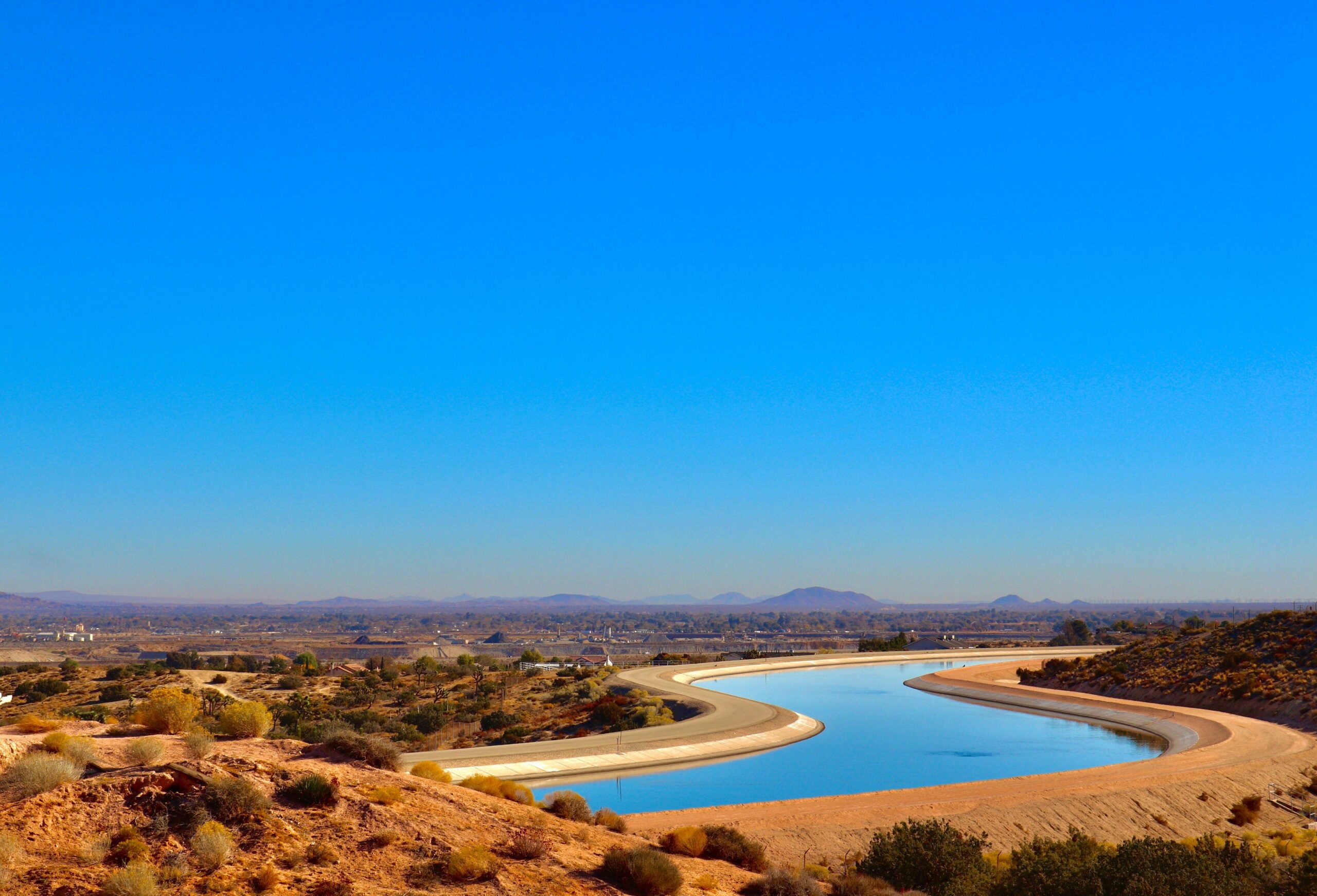A winding water canal flows through arid landscape under a clear blue sky, highlighting the interaction of human infrastructure and natural terrain.